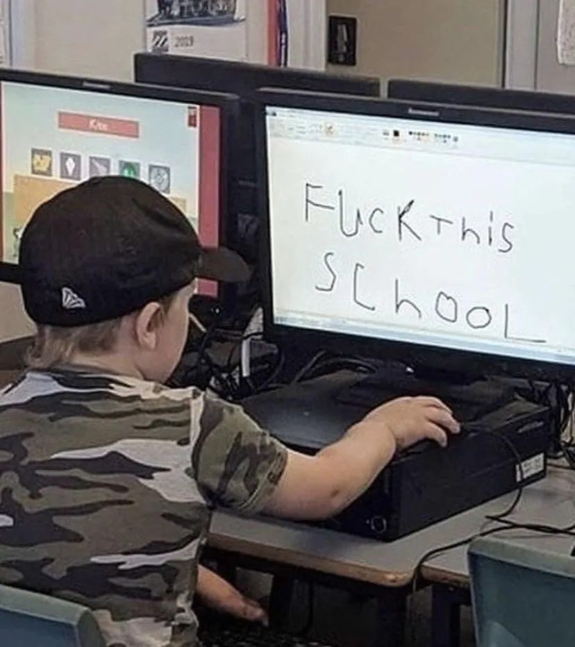 A young child in a camouflage shirt and black baseball cap sits at a school computer desk, facing a monitor displaying the…