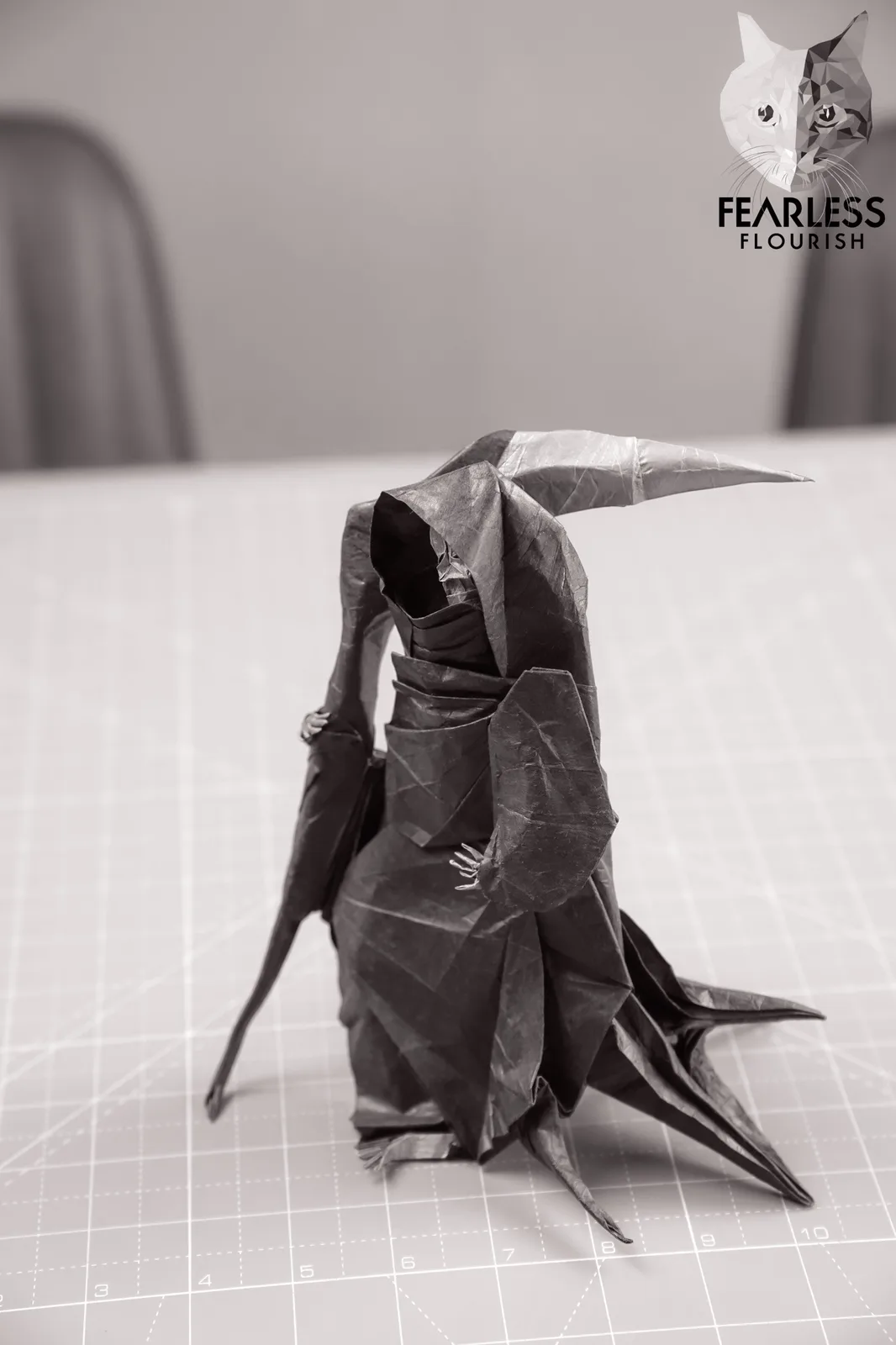 A black-and-white photo shows a detailed origami figure resembling a hooded, winged creature with a long, pointed horn…