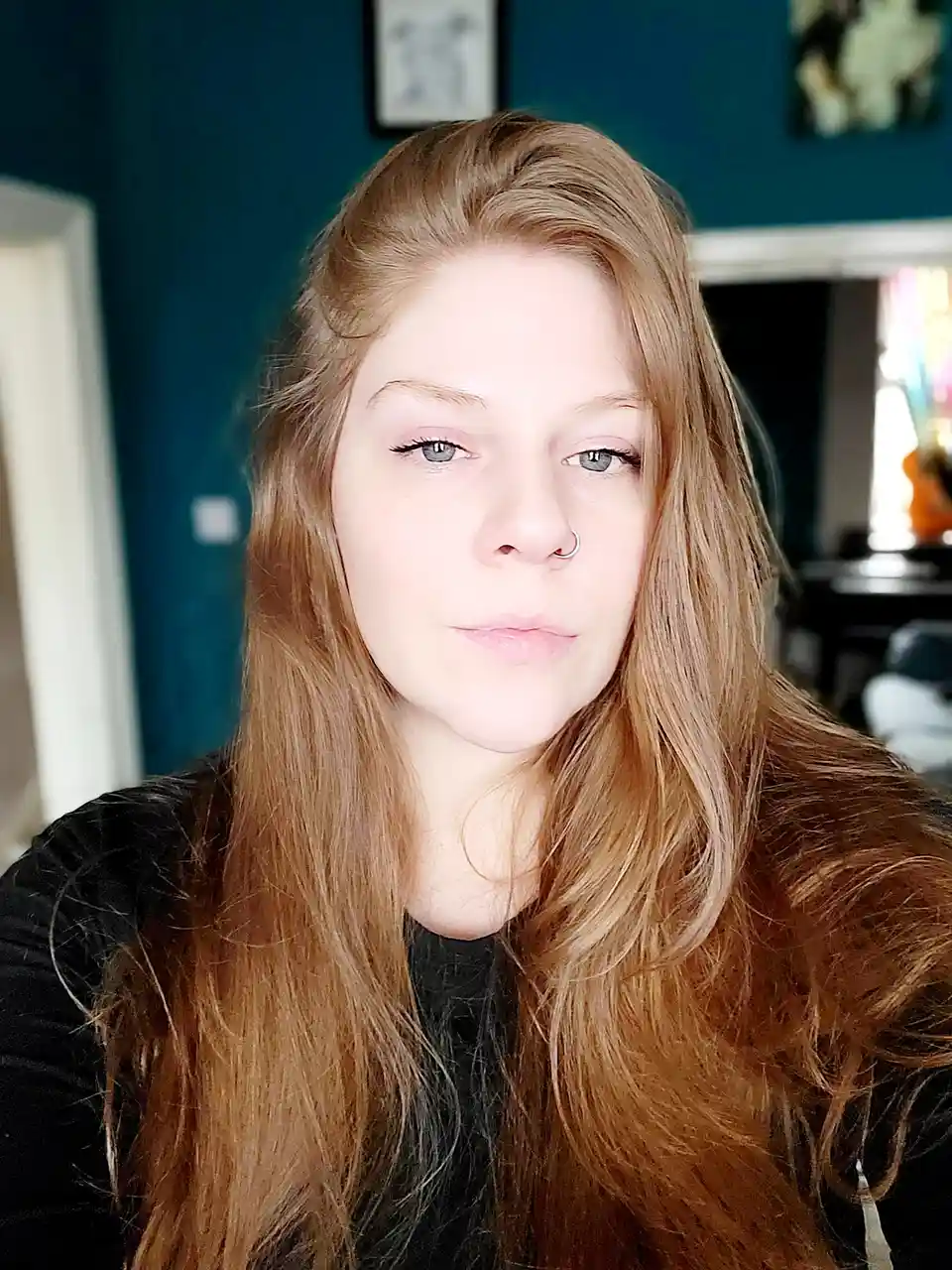 A person with long red hair and a nose ring stares directly at the camera with a neutral expression, wearing a black top…