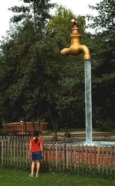 A young girl in a red shirt and denim shorts stands on grass, looking up at a giant golden outdoor water faucet that is…