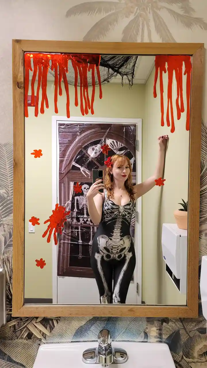 A woman in a full-body skeleton-print bodysuit takes a mirror selfie in a bathroom decorated for Halloween. The mirror has…
