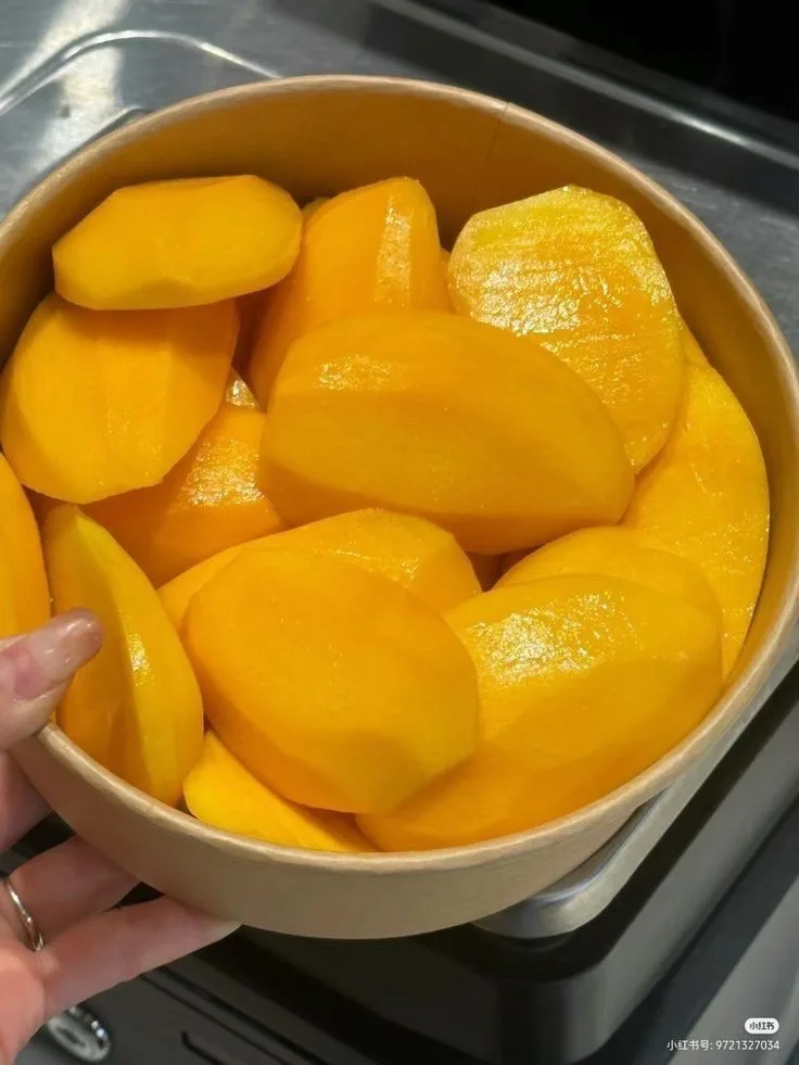 a hand holds a light brown paper bowl filled with bright yellow, peeled, and sliced mango pieces, set against a stainless…
