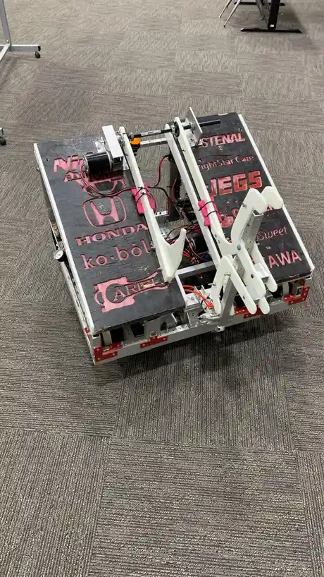 A custom-built robot with a black top plate and white mechanical arms sits on a gray carpeted floor. The plate has red text…
