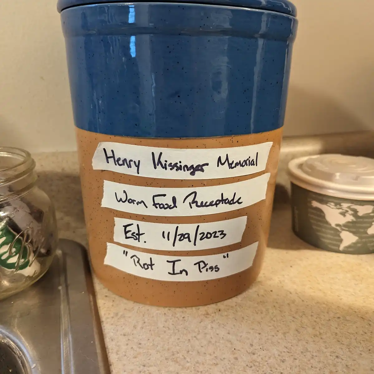A blue and brown ceramic kitchen canister sits on a countertop with four handwritten labels taped to it. The labels read…