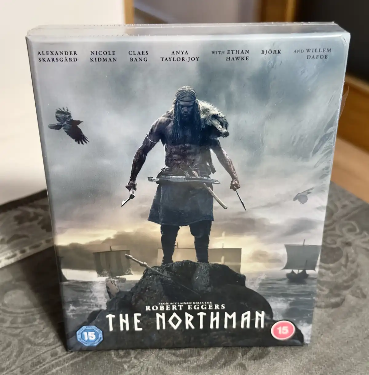 A physical DVD or Blu-ray box set for the movie 'The Northman' is shown standing upright on a patterned surface. The cover…
