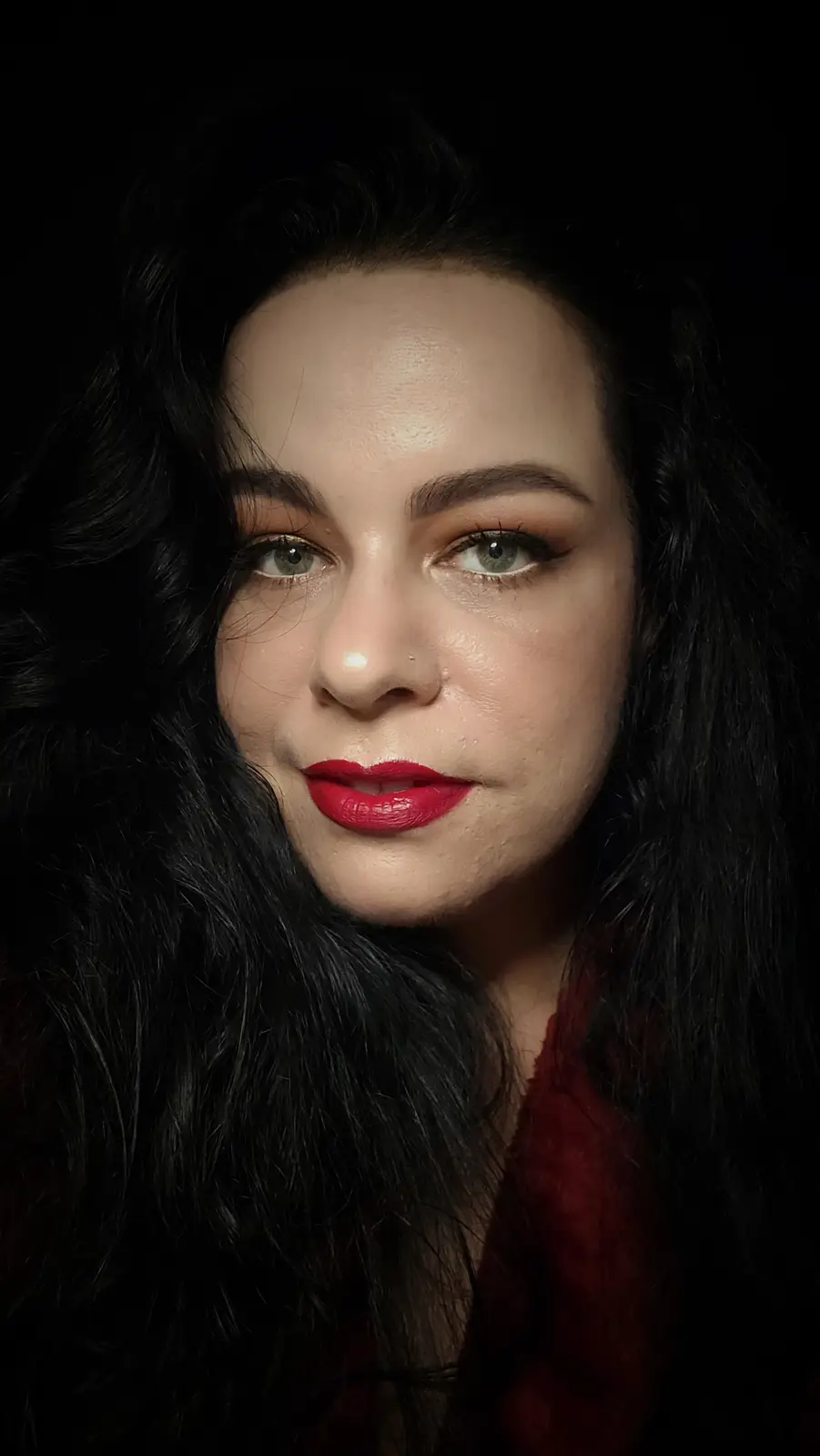 A close-up portrait of a woman with long, dark, wavy hair, green eyes, and bold red lipstick, set against a black…