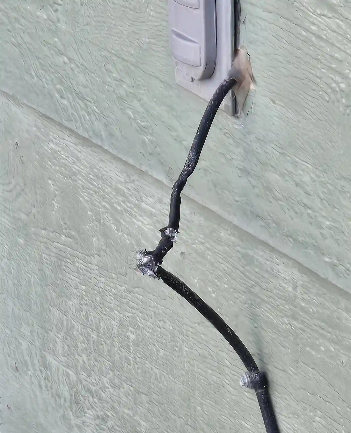 A close-up photo shows a black electrical cord plugged into an outdoor outlet on a light green textured wall. The cord is…