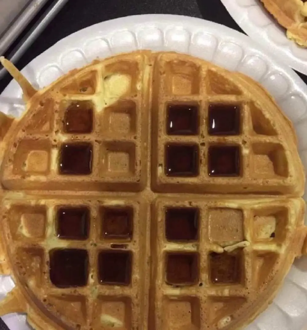 A close-up photo of a golden-brown waffle on a white paper plate, with syrup pooled in some of its square pockets. The…