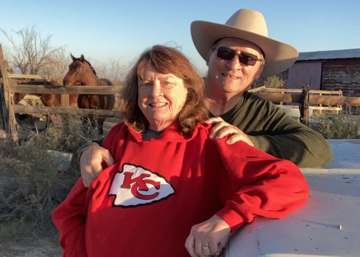 An older couple poses outdoors on a ranch with horses in the background. The woman wears a red Kansas City Chiefs hoodie…