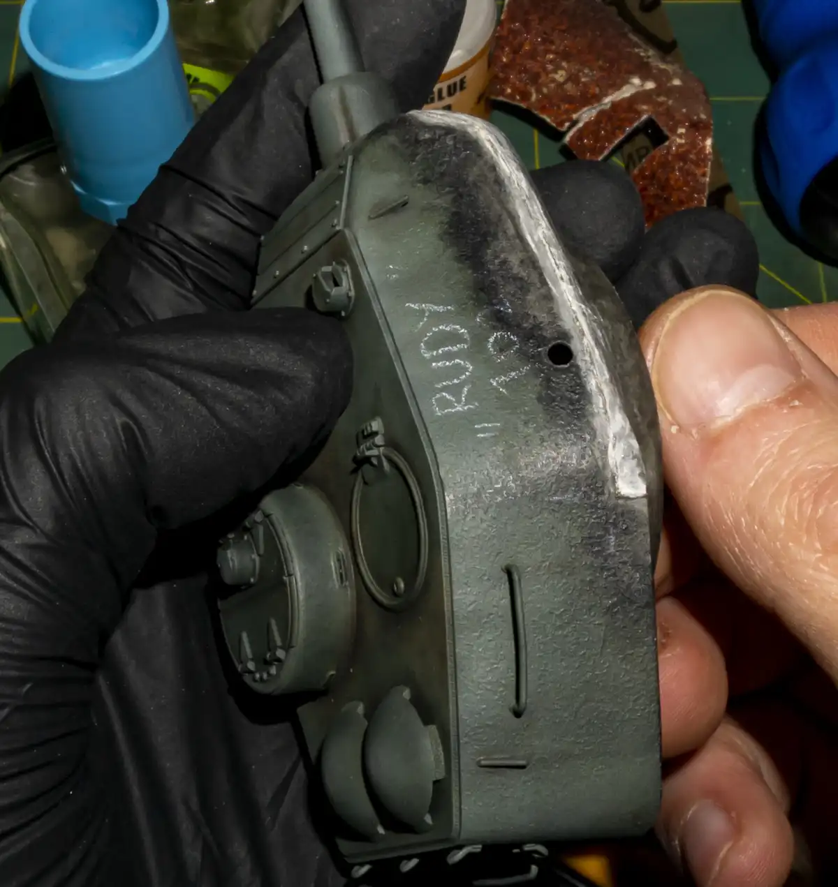 A close-up shot shows a person wearing a black glove holding a small, green-painted military tank model. The side of the…