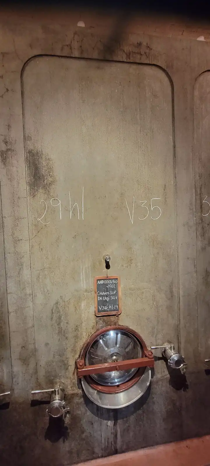 A close-up of a large, weathered concrete wine tank with handwritten markings '29hl V35' on its surface. Below, a small…