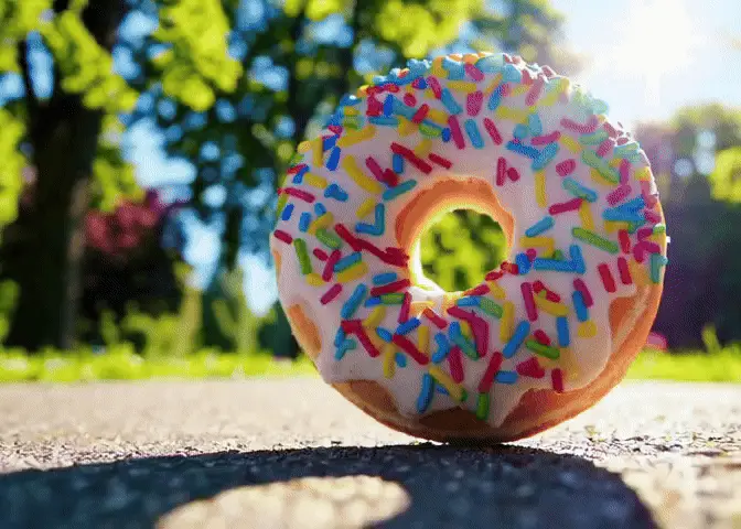 A sequence of six images shows a single, colorful sprinkle-covered donut on a paved path in a sunny park. The donut appears…
