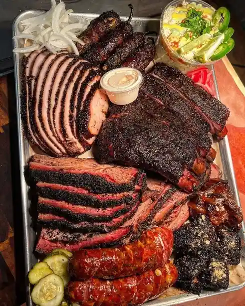 a metal tray overflowing with various smoked meats including sliced brisket, ribs, sausage, and jalapeño poppers…