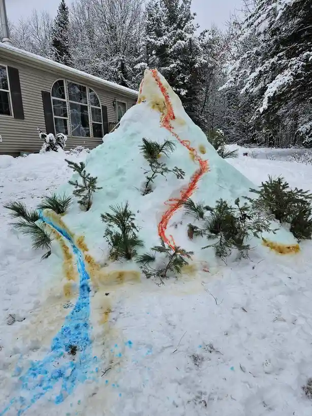 A large, cone-shaped snow mound in a snowy yard, resembling a volcano, is decorated with orange and blue dye streaks flowing…