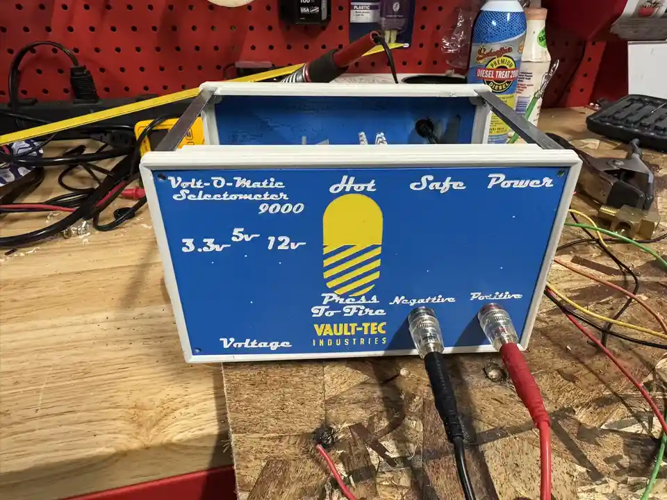 A homemade blue and white box labeled 'Volt-O-Matic Selectometer 9000' with a yellow button and voltage options, mimicking a…