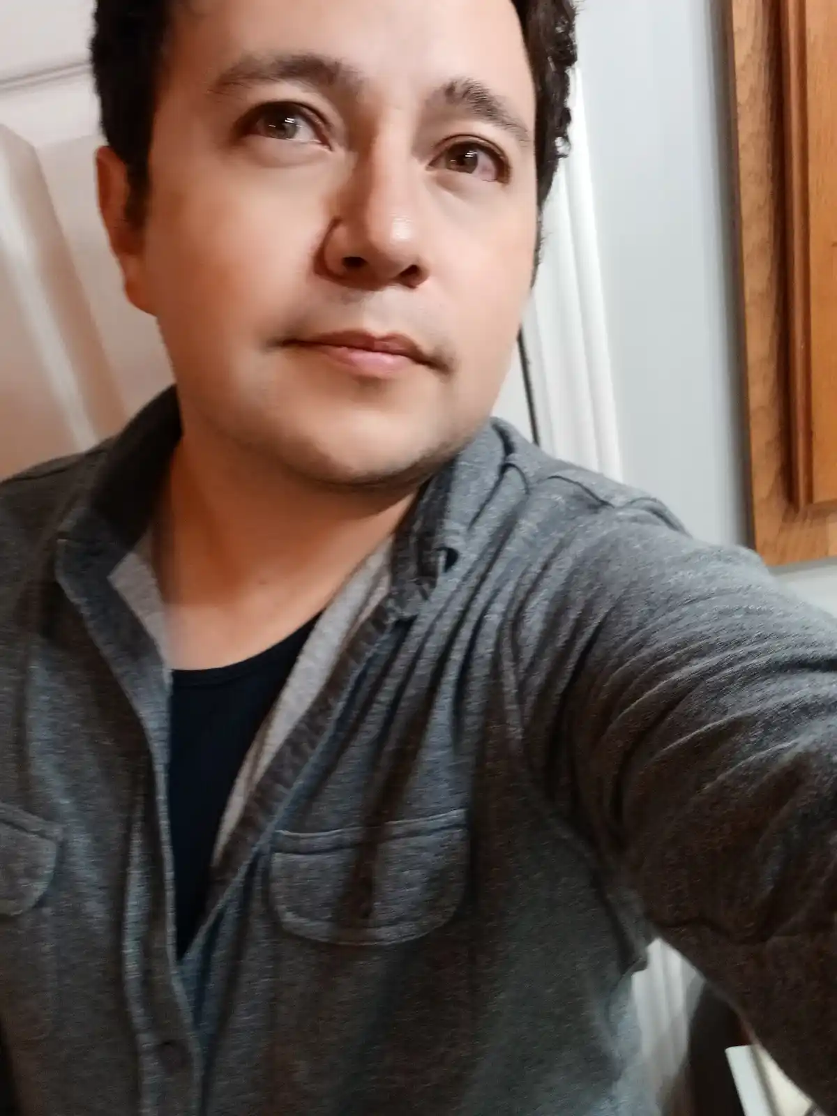 A man in a gray button-up shirt over a black t-shirt takes a selfie indoors, looking upward with a slightly blank or…