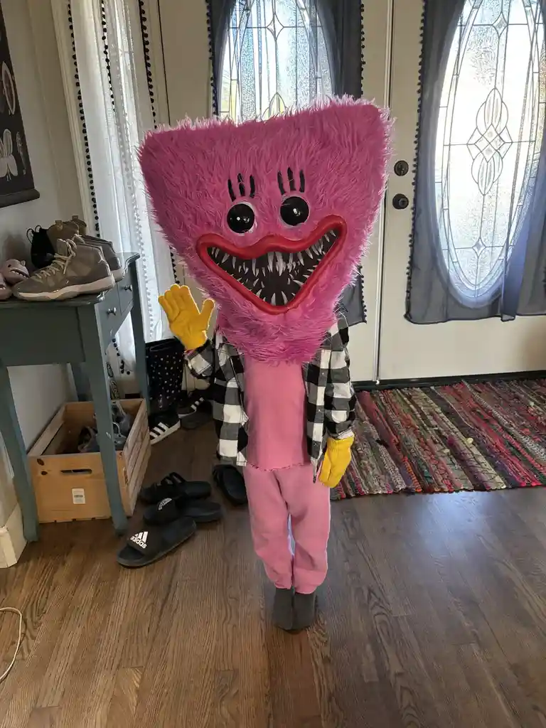 A person wearing a large, fuzzy pink Huggy Wuggy costume with sharp teeth and black eyes stands indoors, waving with a…