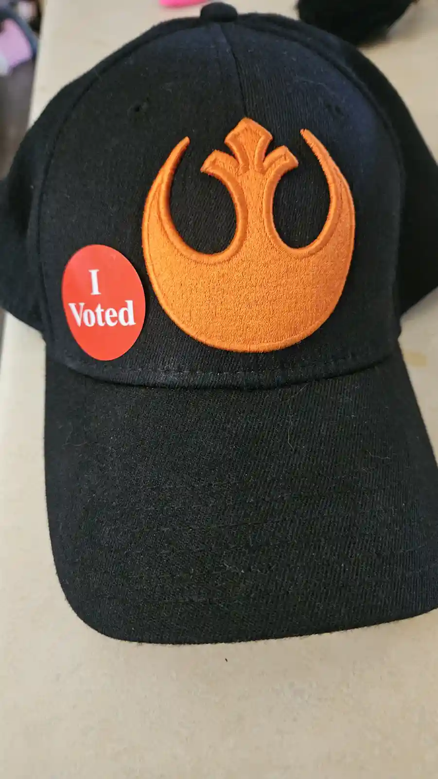 A black baseball cap features a large orange embroidered Rebel Alliance symbol from Star Wars, with a red circular 'I Voted'…