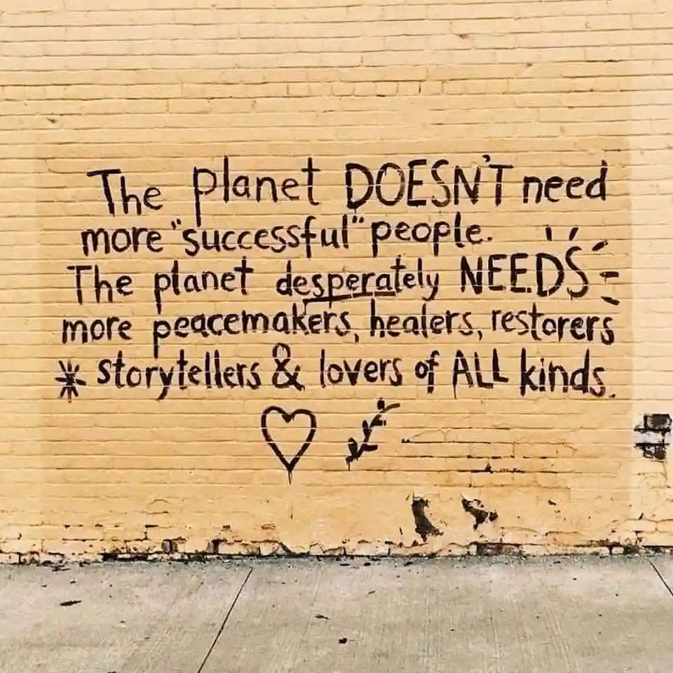 A yellow brick wall has black spray-painted text stating the planet doesn't need more 'successful' people but desperately…