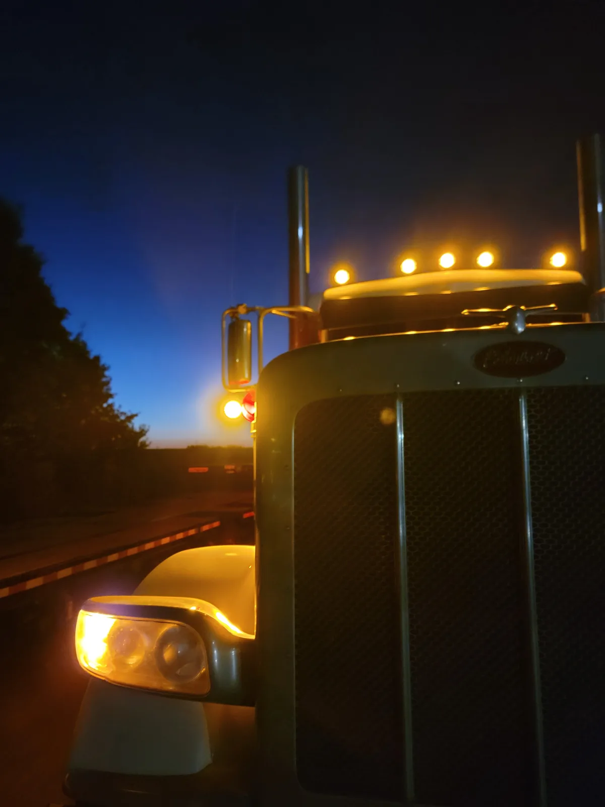 A close-up of a classic Peterbilt semi-truck's front grille and headlights at dusk, with its amber roof lights and side…