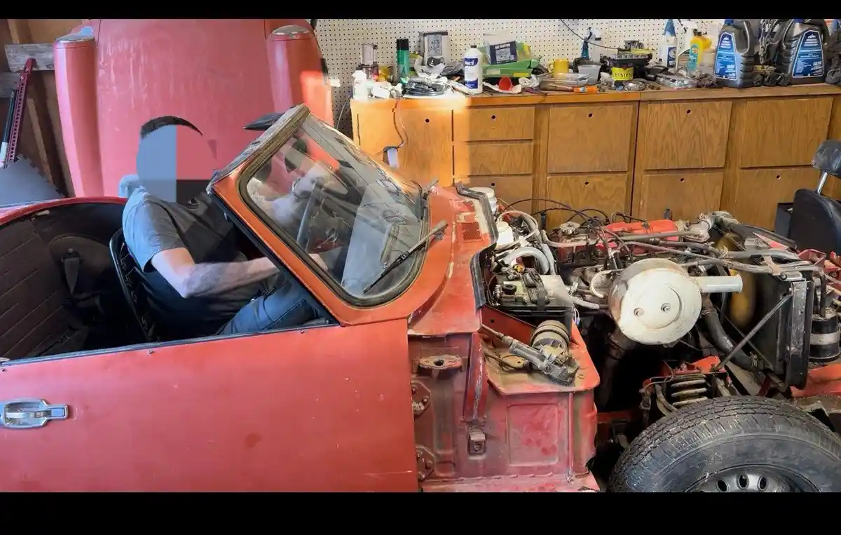 A man sits inside the driver's seat of a red, partially disassembled vintage car in a cluttered garage. The car's hood is…