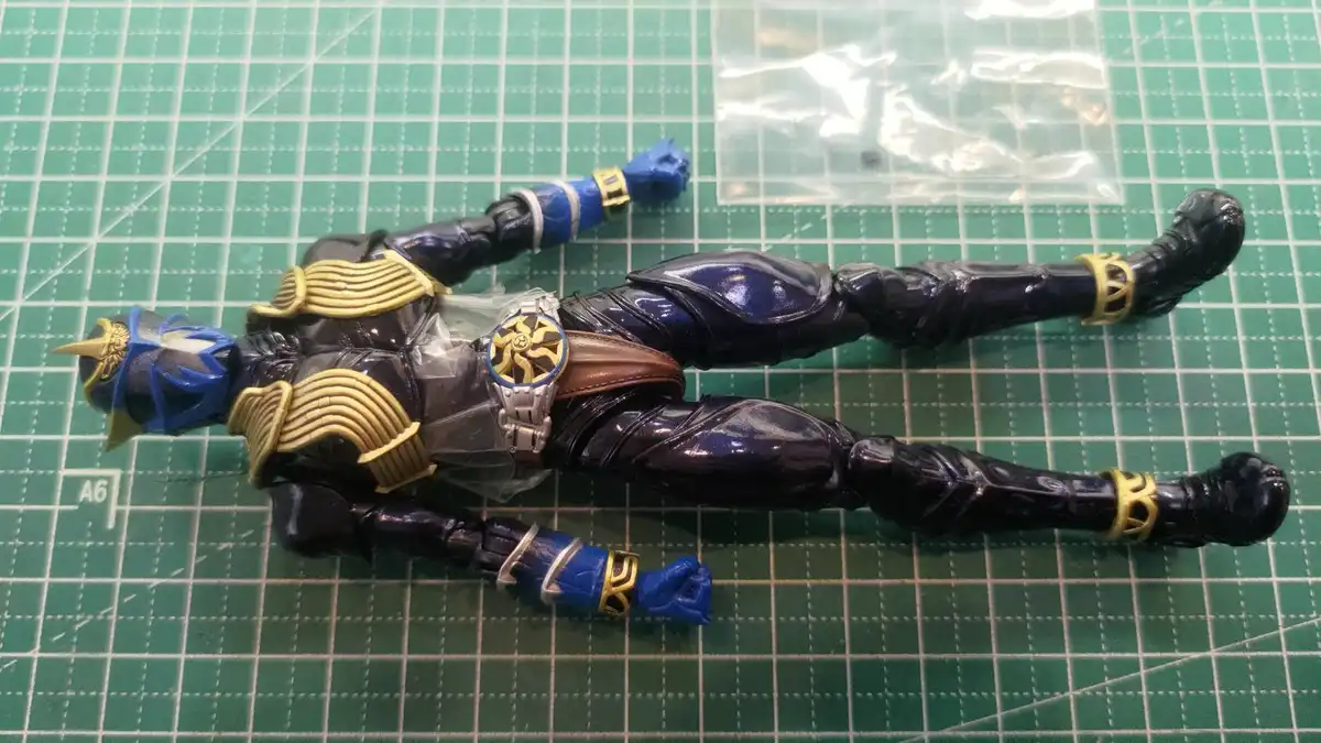 A blue and gold armored action figure, resembling a Kamen Rider, lies flat on its back on a green cutting mat with grid…