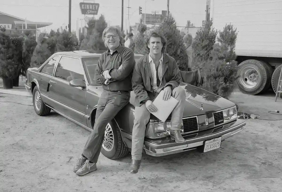 A black-and-white photo shows two men posing beside a 1980s Cadillac sedan. The man on the left, wearing glasses and a…