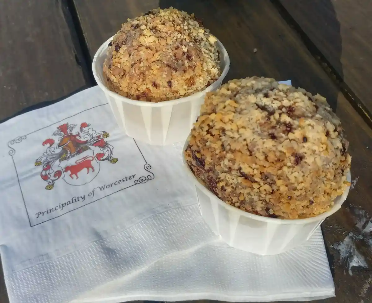 Two muffins with crumbly topping sit in white paper cups on a white napkin printed with a coat of arms and the text…