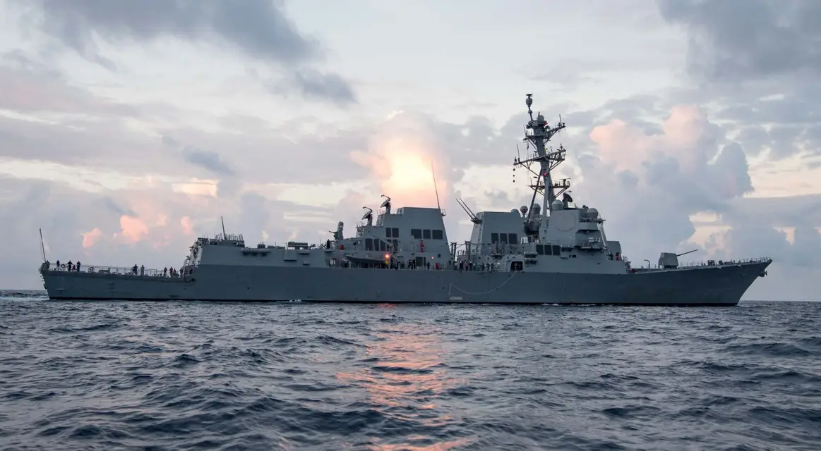 A large gray naval warship sails on the ocean during sunset, with crew members visible on deck. The sky is filled with…