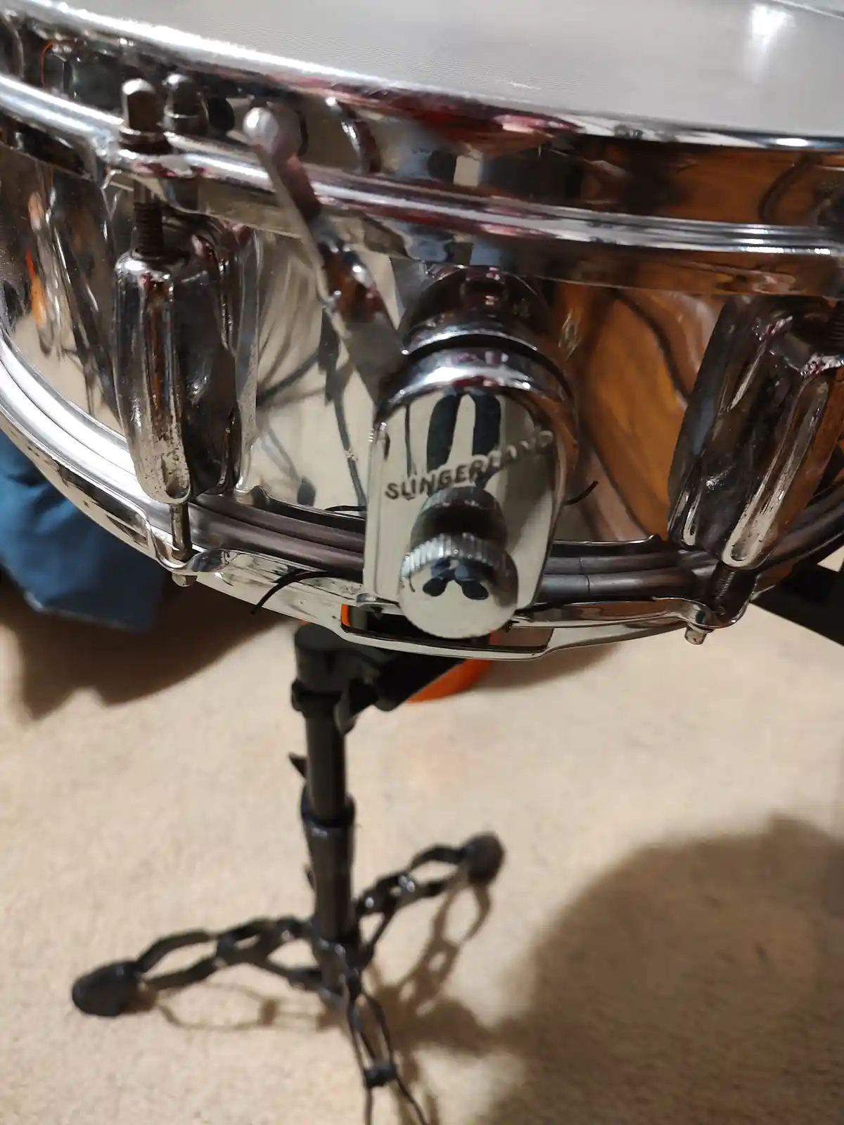 A close-up photo of a shiny chrome snare drum with the brand name 'Slingerland' visible on a tension rod. The drum is…