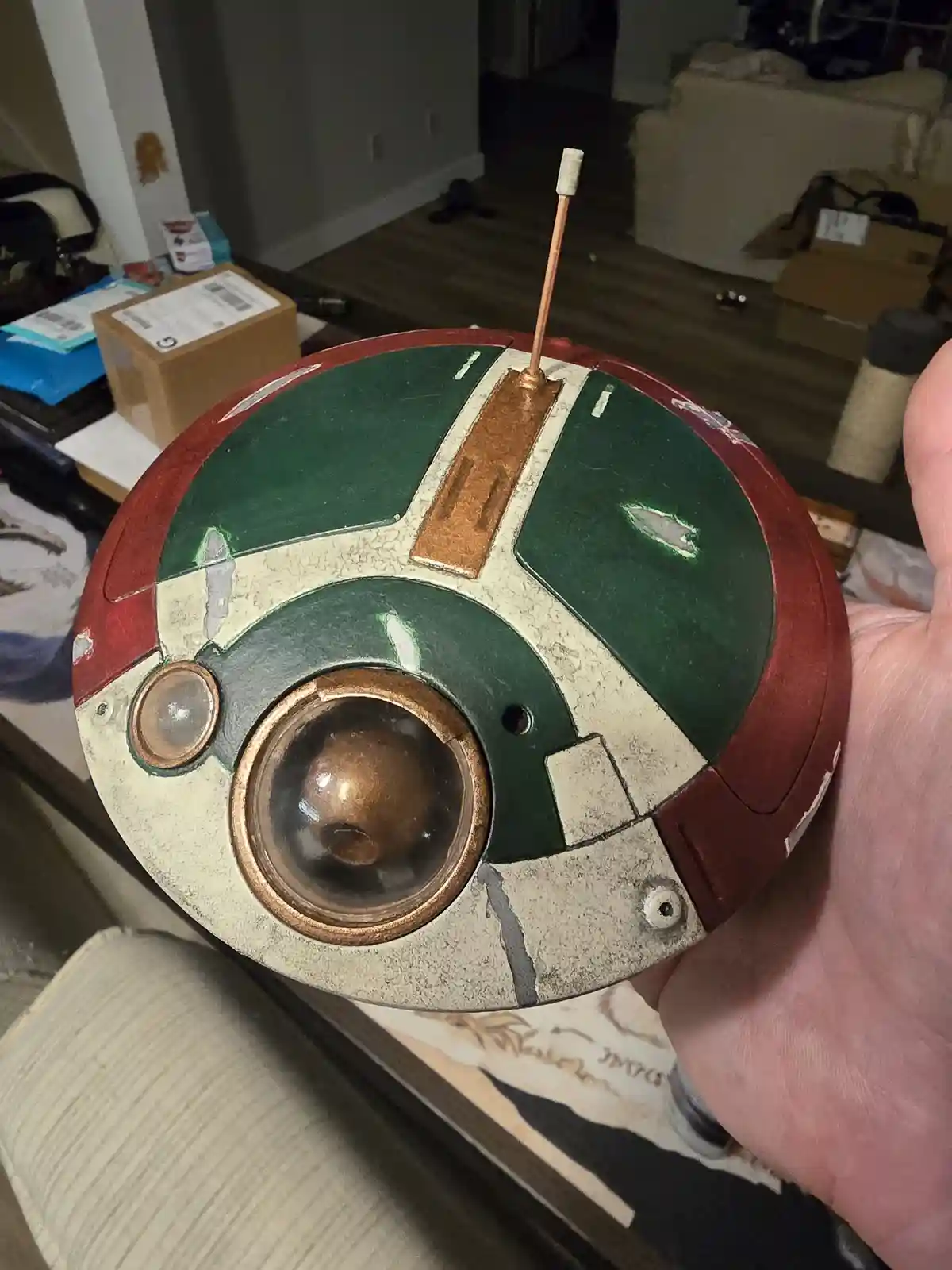 A hand holds a spherical, crudely painted helmet resembling Boba Fett's iconic armor, featuring green, red, and beige…
