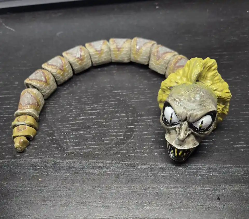 A 3D-printed snake bracelet with a skull-headed head and segmented body lies curved on a dark, scratched surface. The skull…