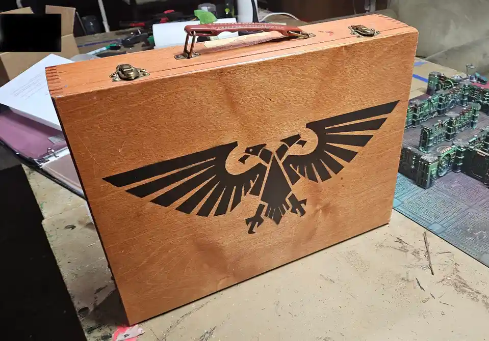 A wooden box with a red leather handle and brass latches sits on a cluttered workbench, featuring a black, stylized eagle…