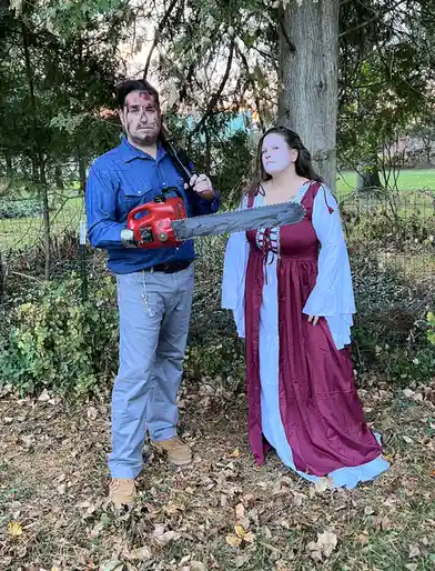 A man with blood makeup and a chainsaw over his shoulder stands next to a woman in a medieval-style dress, both looking at…