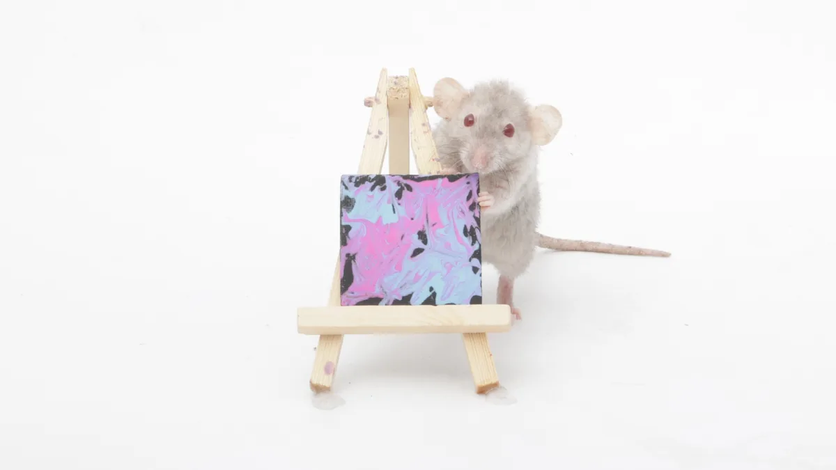 A small gray mouse stands upright on a white surface, holding a miniature wooden easel that displays a colorful abstract…