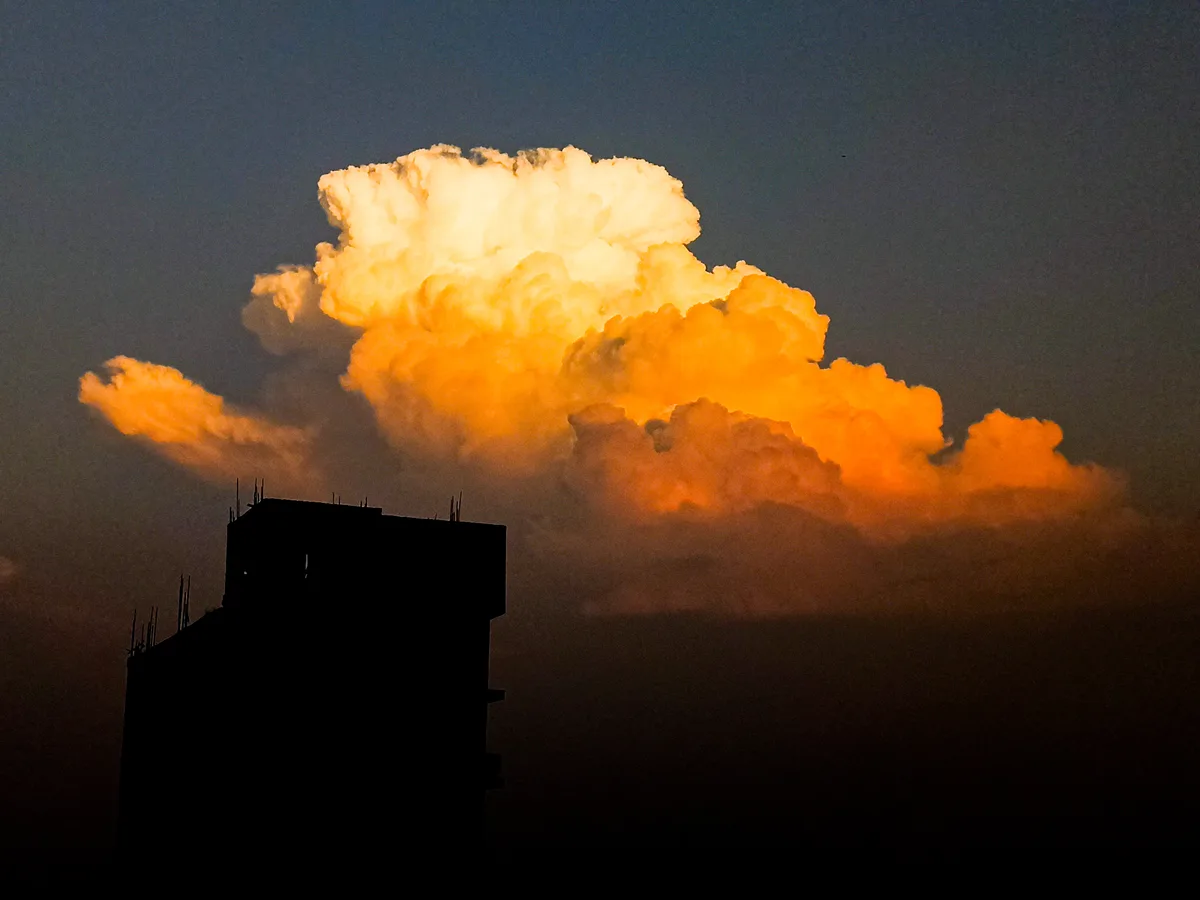 A dramatic sky photo shows a large, fiery-orange cloud illuminated by sunset or sunrise, towering above the dark silhouette…
