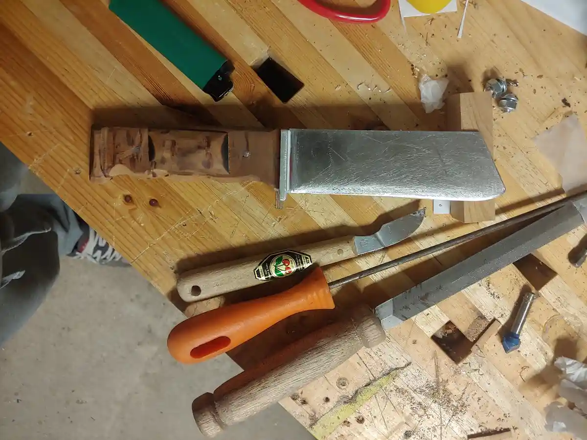 A wooden workbench holds a large, homemade meat cleaver with a wooden handle and metal blade, alongside various woodworking…
