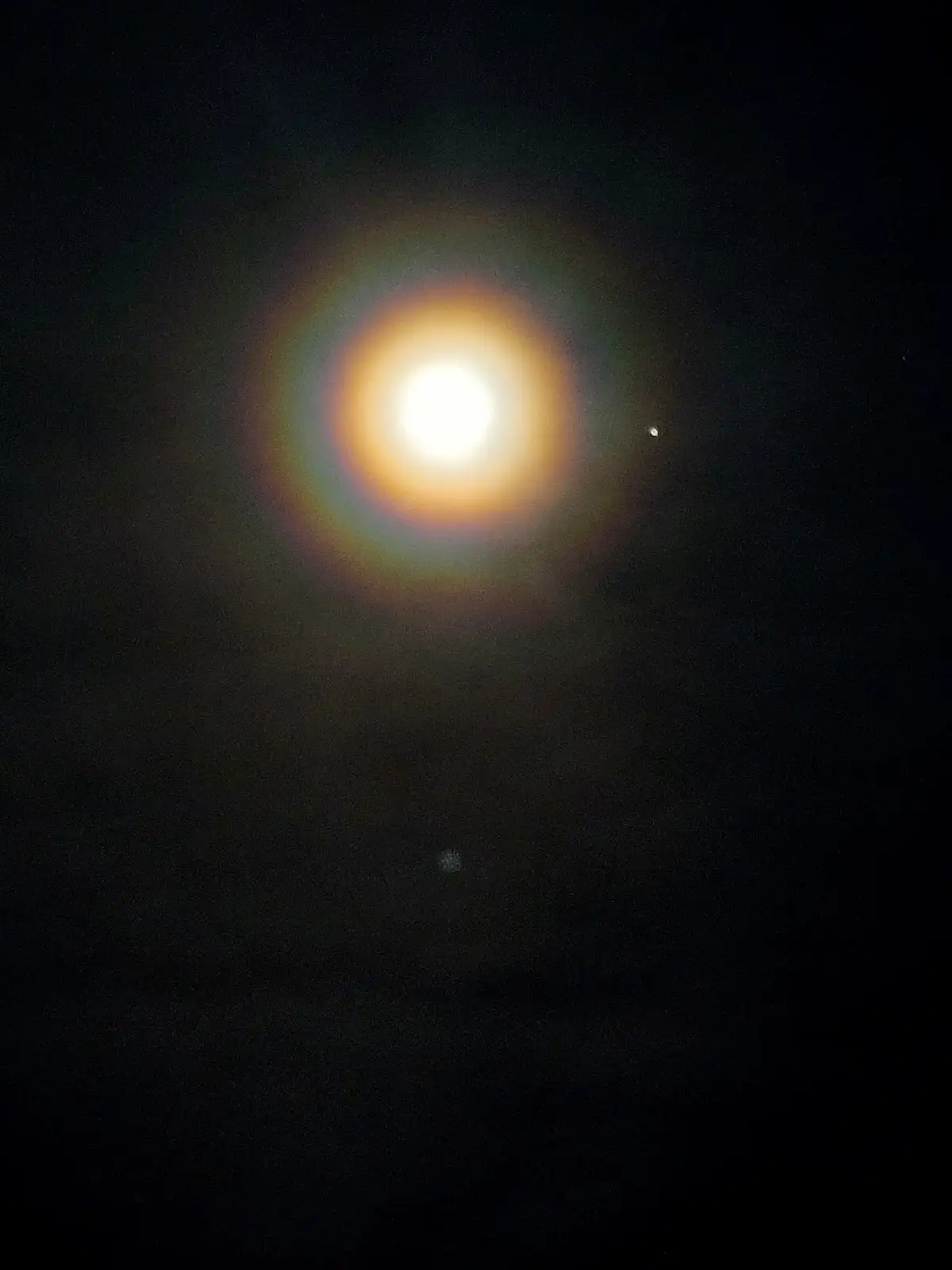 A dark night sky shows a bright, glowing orb with a rainbow halo, resembling the sun or moon, with two small, faint dots…