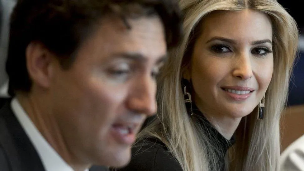A close-up photo shows a man in a suit speaking while looking down and to the side, with a woman with blonde hair and large…