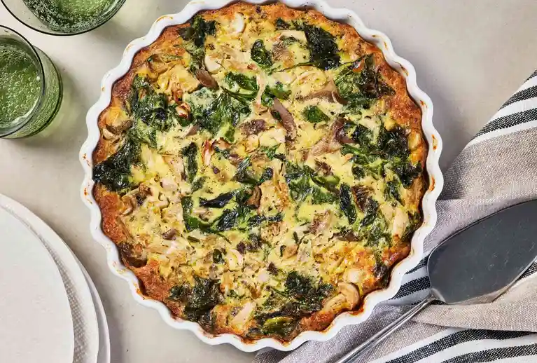 A top-down photo of a baked quiche in a white scalloped pie dish, featuring visible spinach, mushrooms, and chicken. Two…