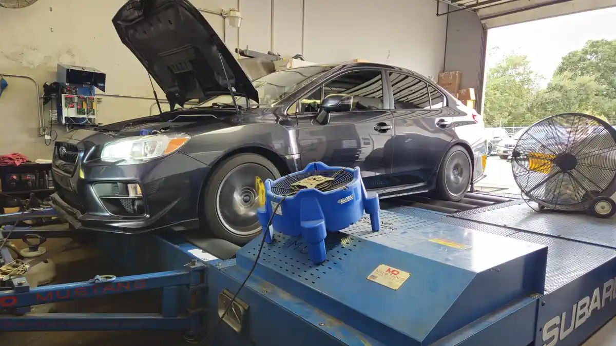 A dark gray Subaru WRX is positioned on a blue Mustang Dyno dynamometer in a garage, with its hood open. In front of the…