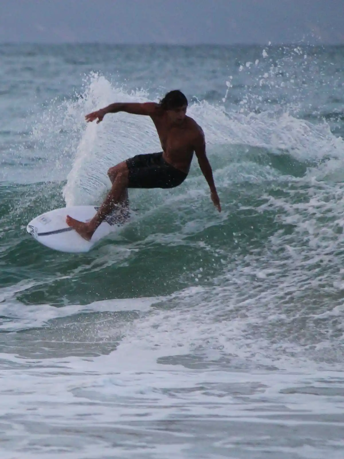 A shirtless man with dark hair is surfing on a white surfboard, leaning into a turn on a greenish wave with white foam…