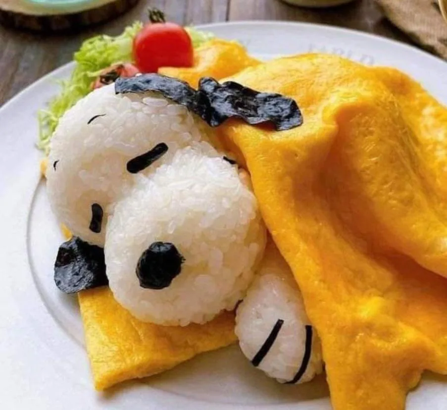 A plate of food features a rice ball shaped like Snoopy, made with white rice and black nori seaweed for facial features and…