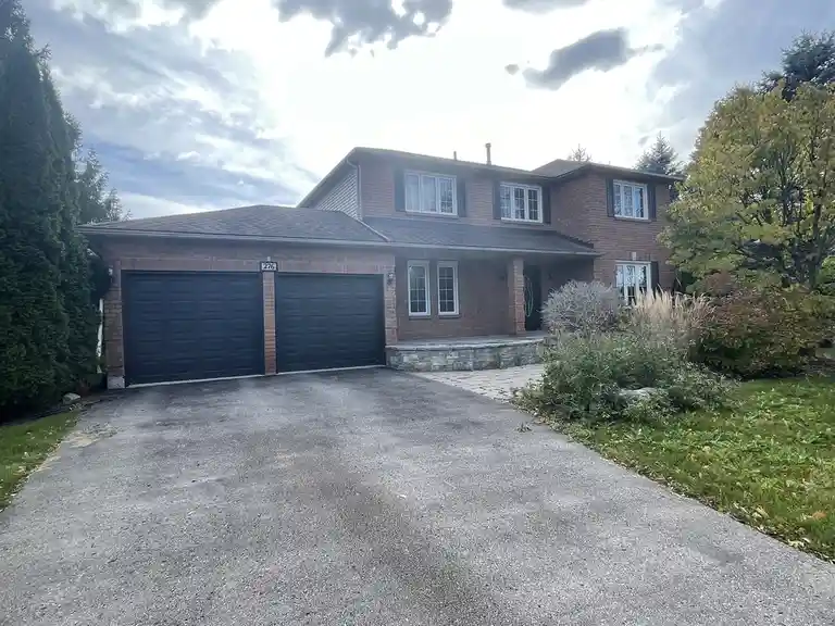 A photo of a two-story brick house with a two-car garage, driveway, and surrounding trees and grass under a cloudy sky. The…
