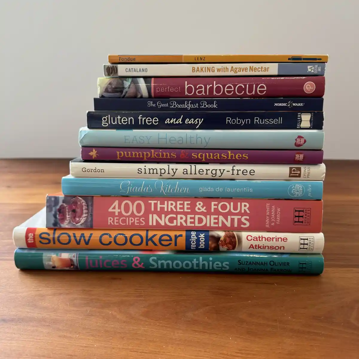 A tall stack of various cookbooks sits on a wooden table, featuring titles like 'Fondue', 'BAKING with Agave Nectar'…