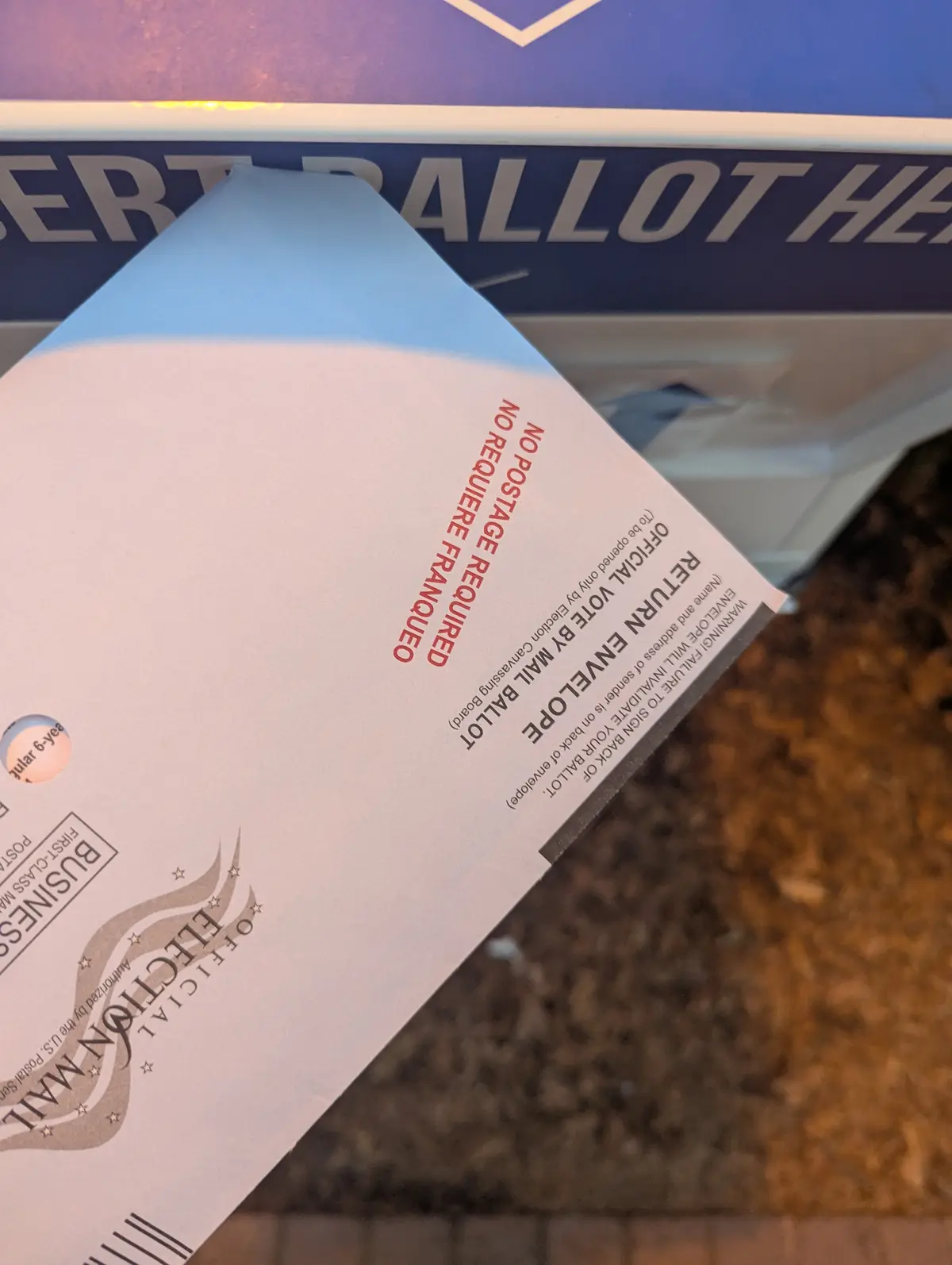 A close-up photo shows a white official election mail ballot return envelope with red text stating 'NO POSTAGE REQUIRED' and…