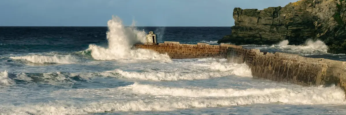 A wide shot of a stormy ocean with large waves crashing against a weathered stone pier and cliff. A small, dilapidated stone…