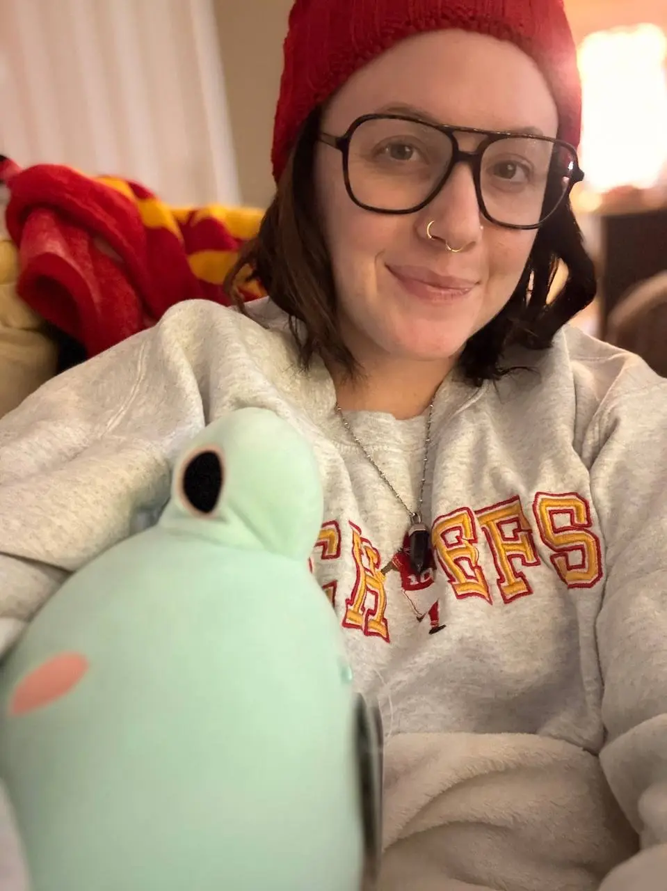 A person wearing a red beanie, large black-rimmed glasses, and a grey 'CHIEFS' sweatshirt smiles while holding a light green…