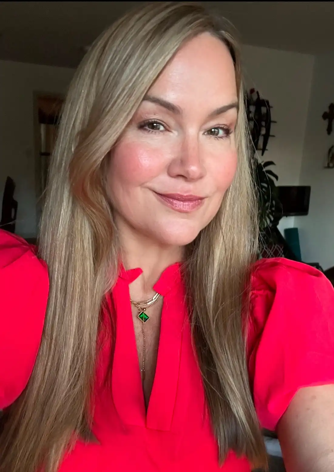 A woman with long blonde hair and a bright red blouse smiles at the camera, wearing a necklace with a green pendant. The…