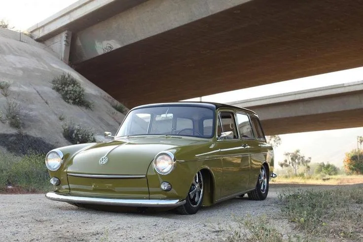 A vintage olive-green Volkswagen station wagon with custom lowered suspension and modern wheels is parked under a concrete…